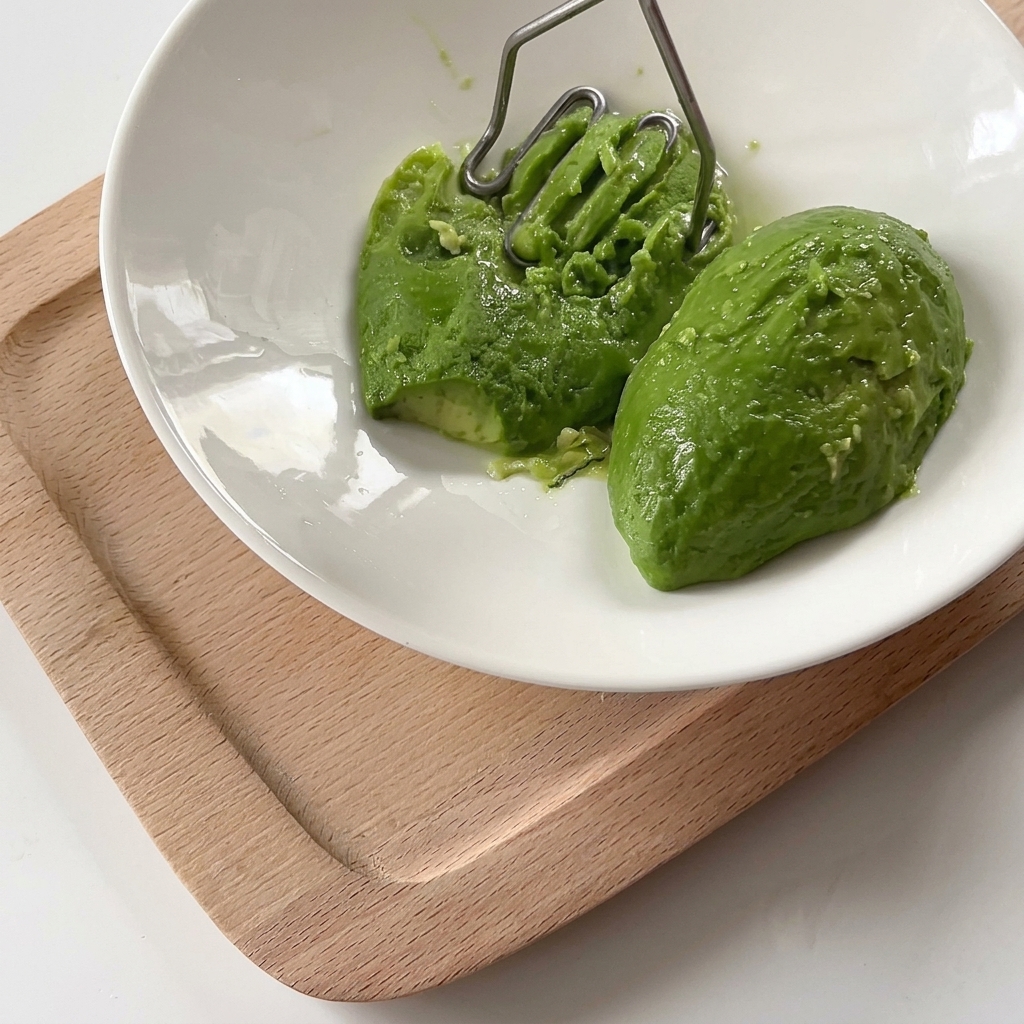 A metal potato masher pressing into two vibrant green avocado halves inside a white ceramic bowl to create a smooth mash.
