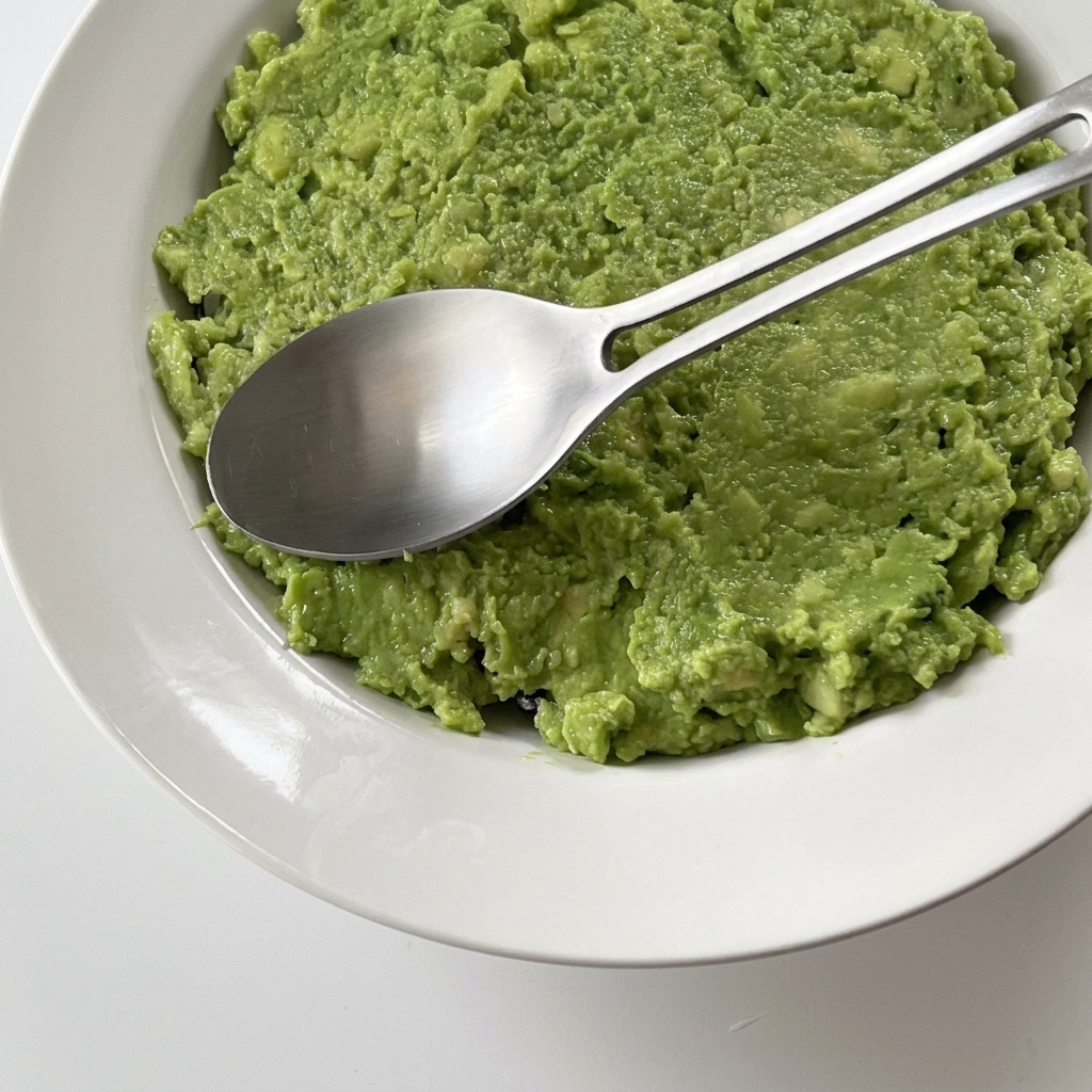 A close-up shot of bright green mashed avocado being spread smoothly into a white bowl using a metal spoon.