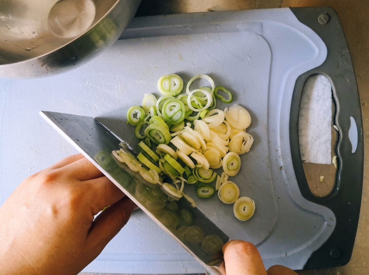A chef using a large kitchen knife to slice fresh green onions into thin rounds on a blue cutting board.