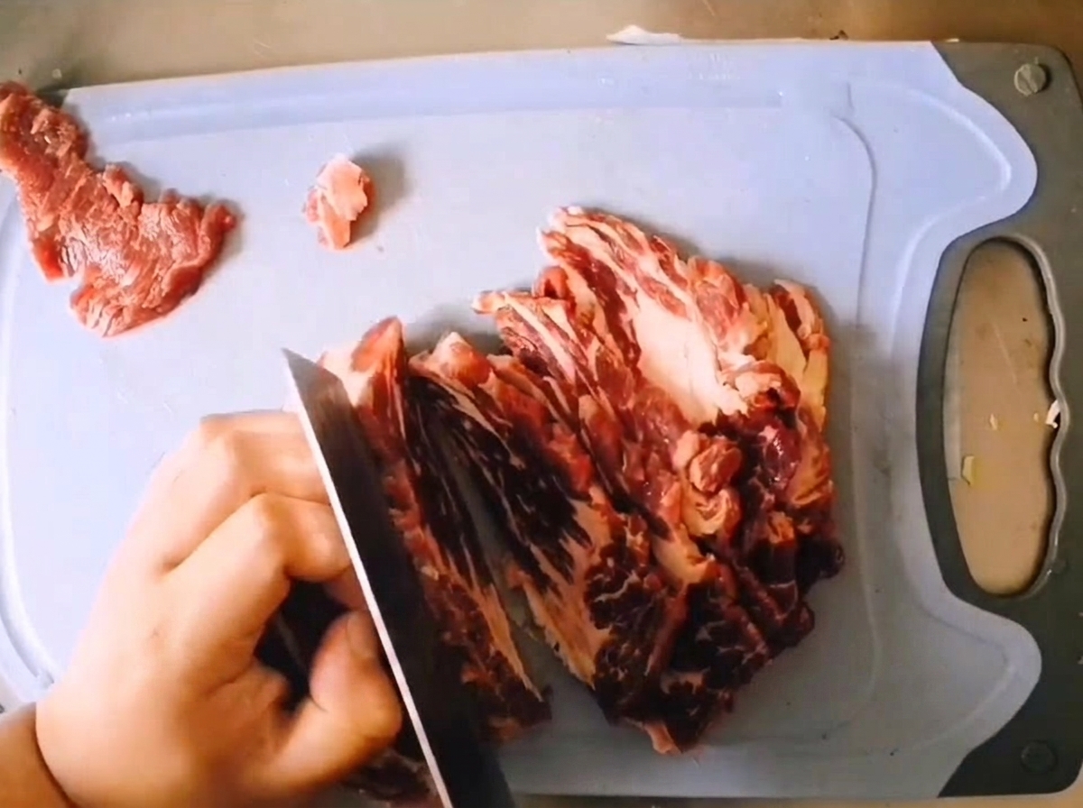 Slicing a piece of raw marbled beef chuck into thick slices with a sharp knife on a light blue cutting board.