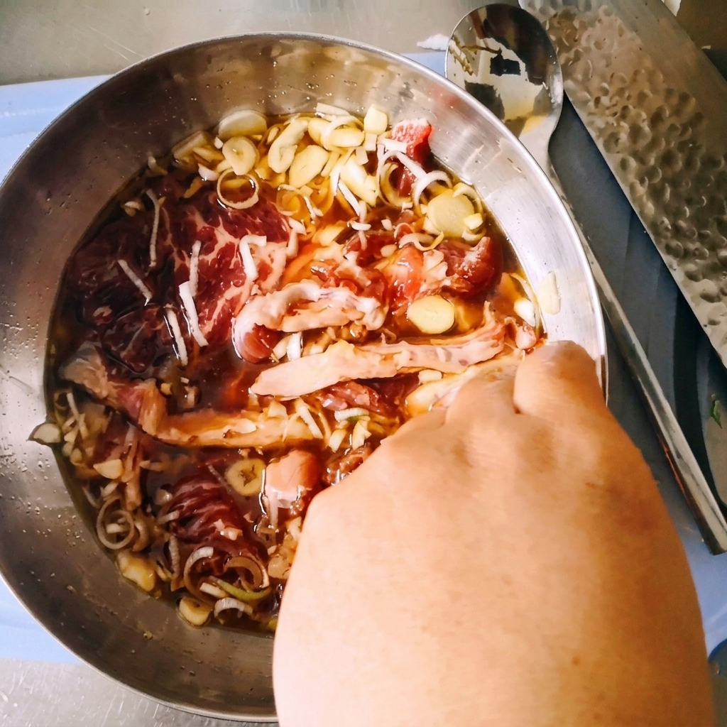 A hand mixing raw sliced beef chuck inside a stainless steel bowl with the dark soy marinade, scallions, and garlic.