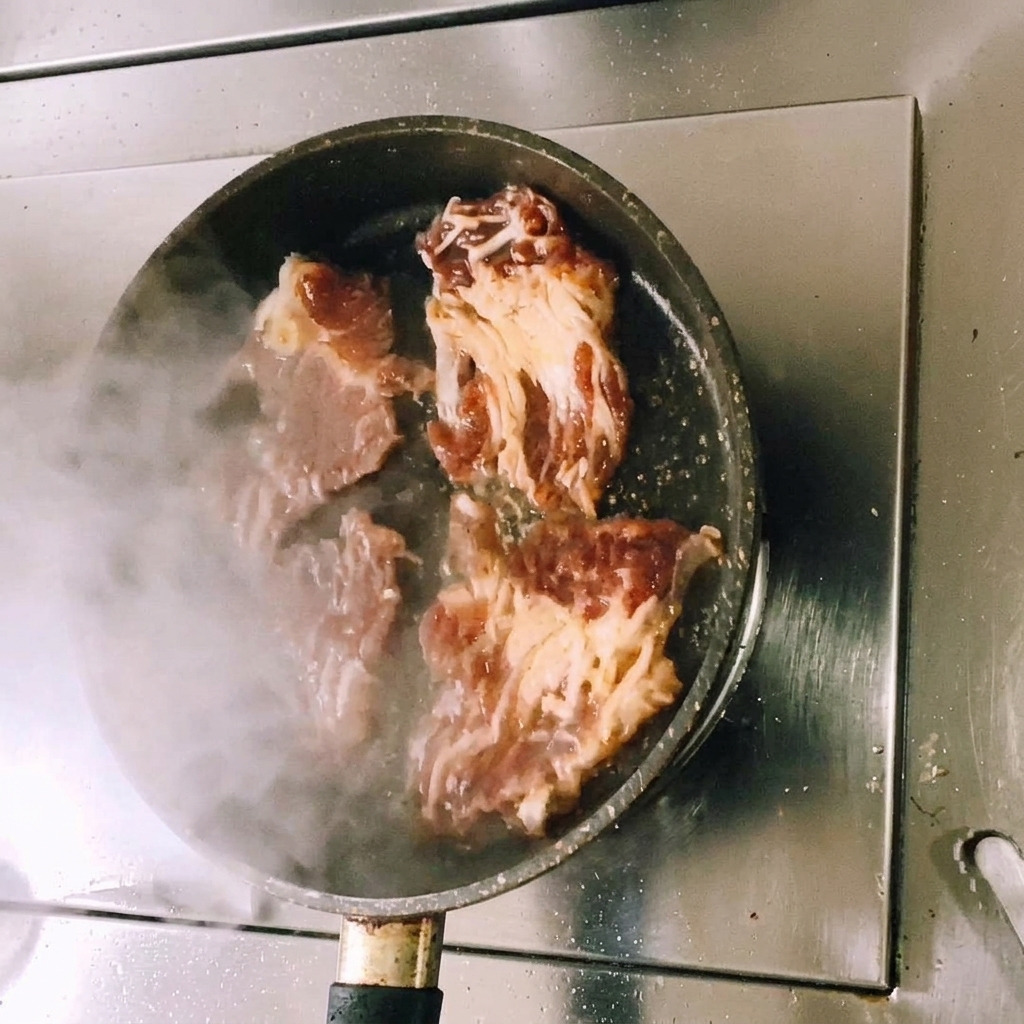 Slices of marinated beef being pan-fried in a flat pan with visible steam rising as the meat begins to brown.