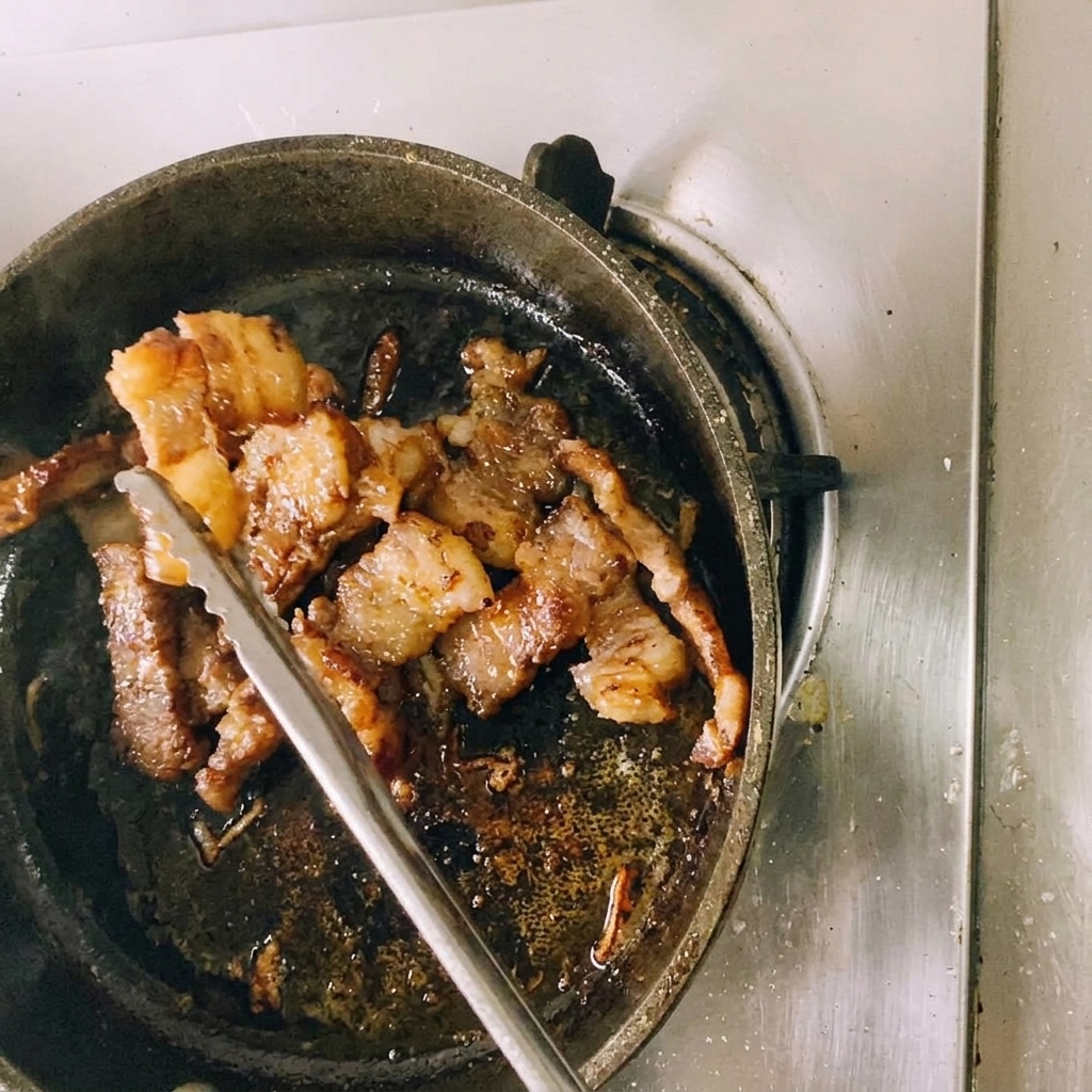 A pair of metal kitchen tongs holding up a piece of seared, browned beef while cutting it into smaller portions over the frying pan.