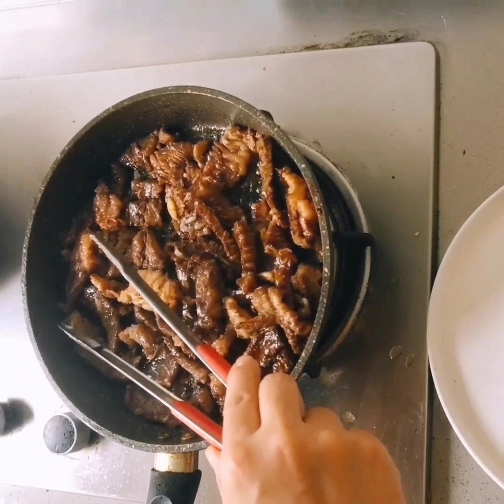 Bite-sized pieces of cooked beef simmering in a rich, dark sauce reduction within a skillet while being tossed with tongs.