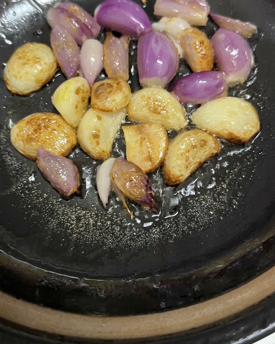 Whole garlic cloves and purple shallots sizzling and lightly browning in oil at the bottom of a pan.