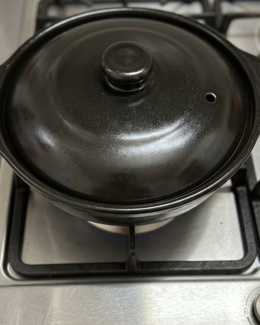 A closed black claypot lid heating over a blue gas stove flame.