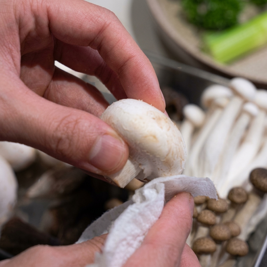 Close up of hands using a white paper towel to gently clean a fresh white button mushroom.