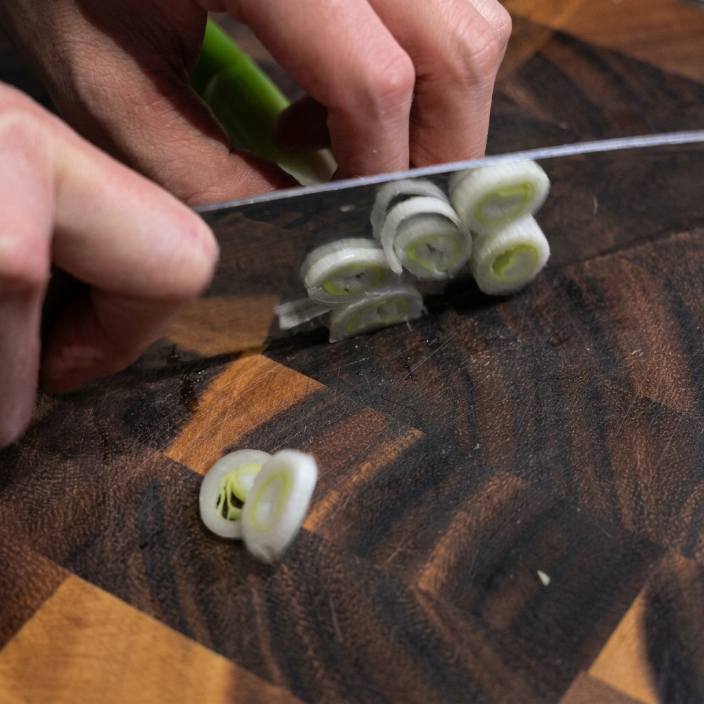 Hands holding a chefs knife slicing a fresh green leek into round rings on a dark wooden cutting board.