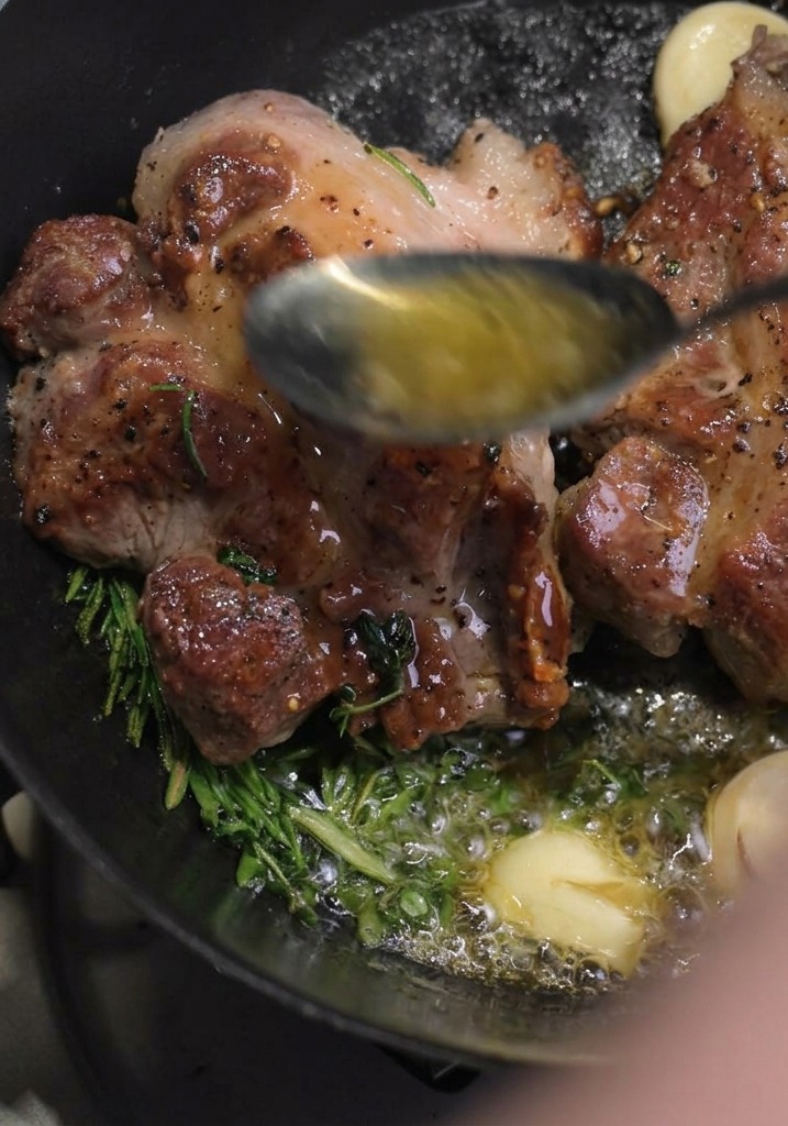 A spoon collecting melted hot butter and pouring it over the surface of seared pork chops cooking with fresh rosemary and garlic.