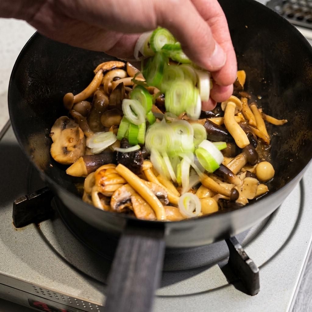 Slices of green leek being added by hand into a pan containing sautéed mushrooms.