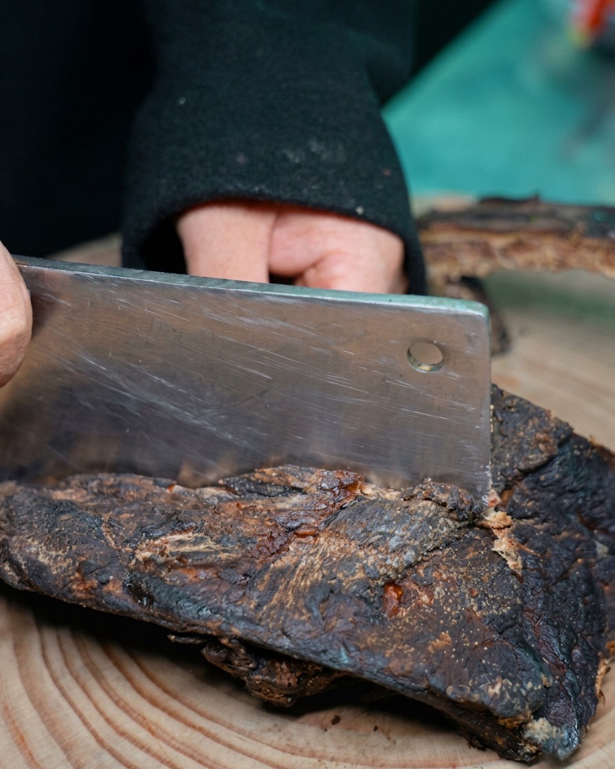 A persons hands using a large metal cleaver to chop a dark slab of cured pork ribs on a wooden cutting board.