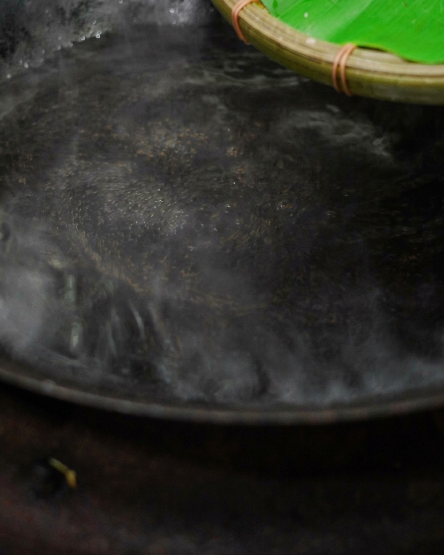 Chopped cured ribs being poured out from a woven bamboo basket into a steaming wok filled with boiling water.