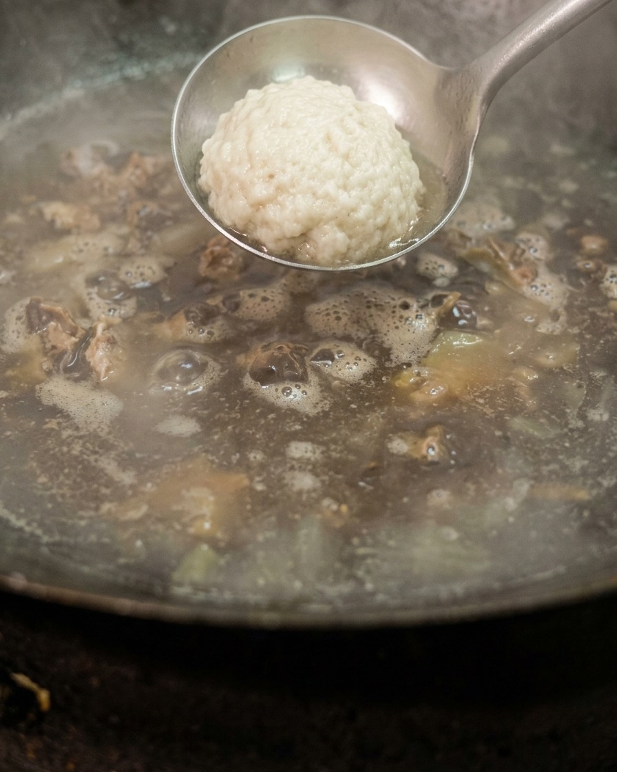 A metal ladle holding a clump of white salt being lowered into a wok of boiling liquid containing pieces of cured meat.
