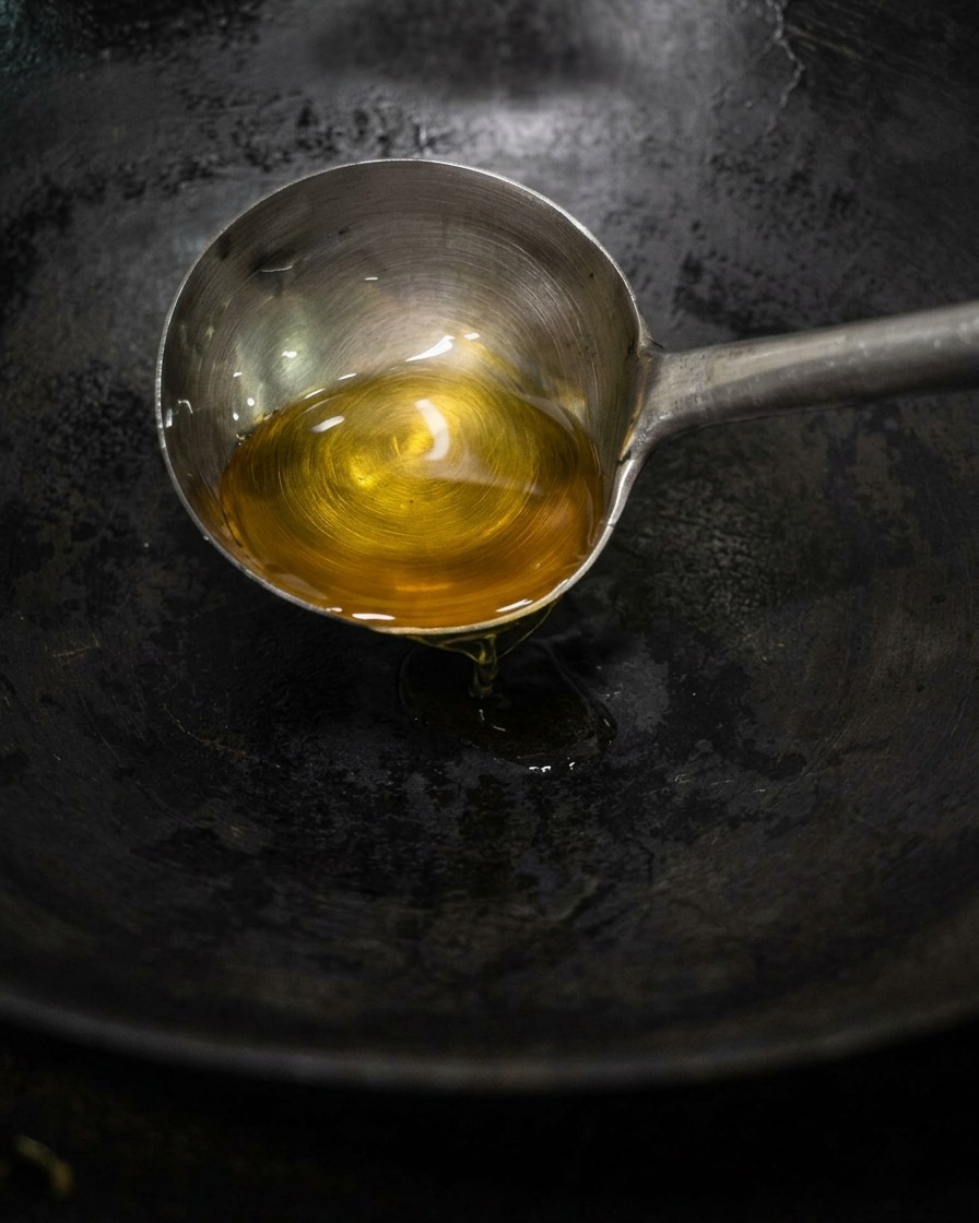 A metal ladle pouring golden tea oil into a dark, heated wok.