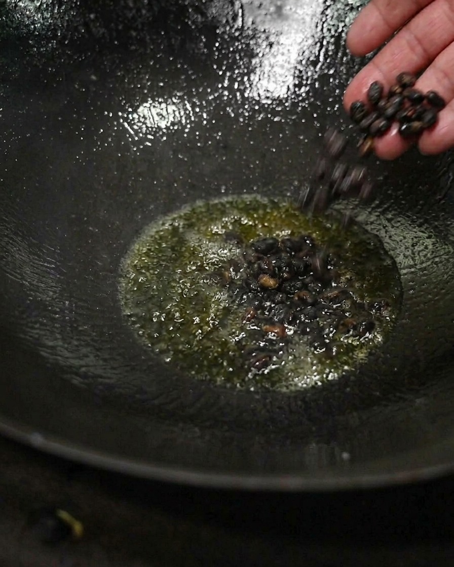 A hand dropping a small cluster of dark fermented black beans into glistening hot tea oil at the bottom of a dark wok.