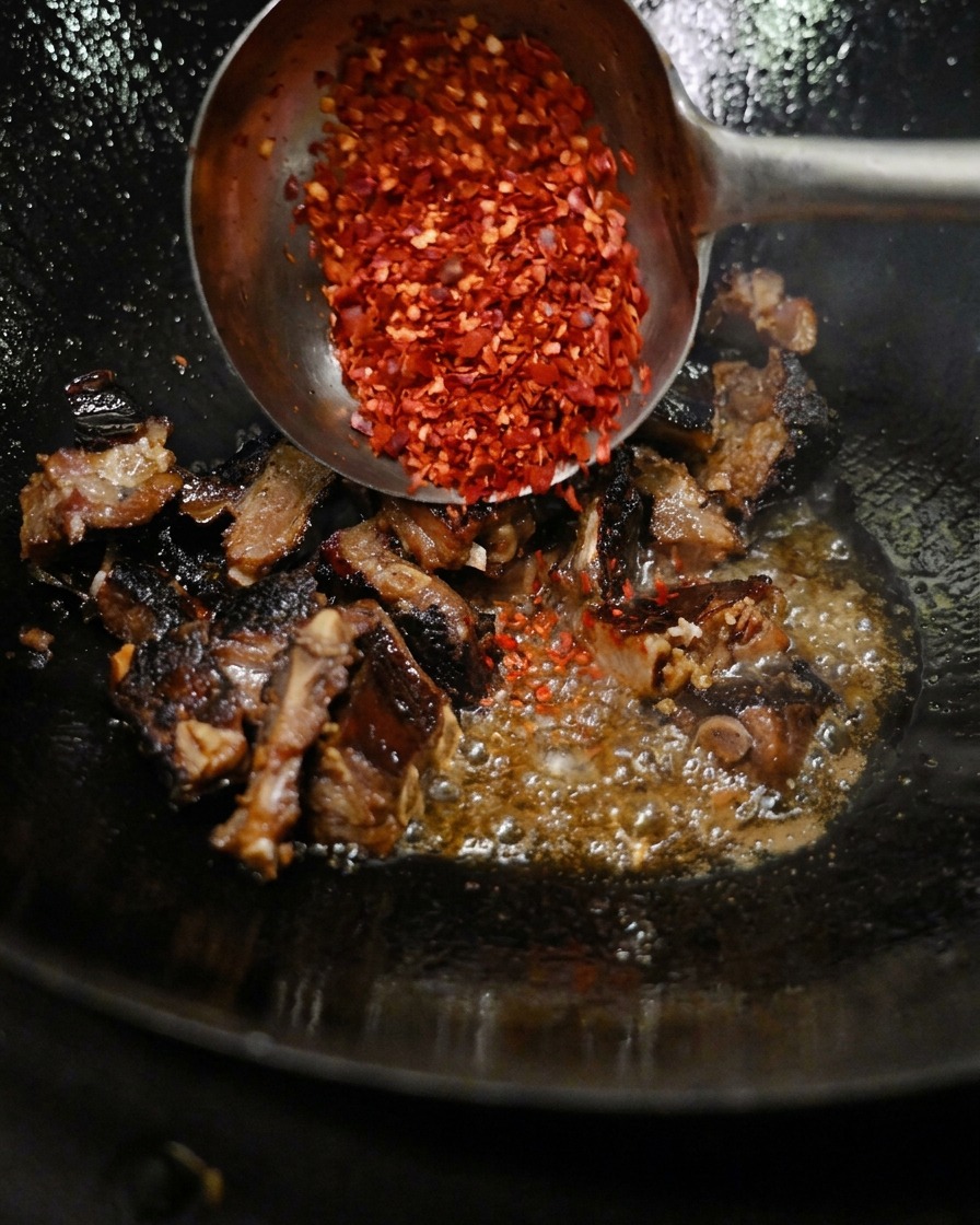A metal ladle pouring a pile of ground red chili flakes over seasoned pork ribs stir-frying in a hot wok.