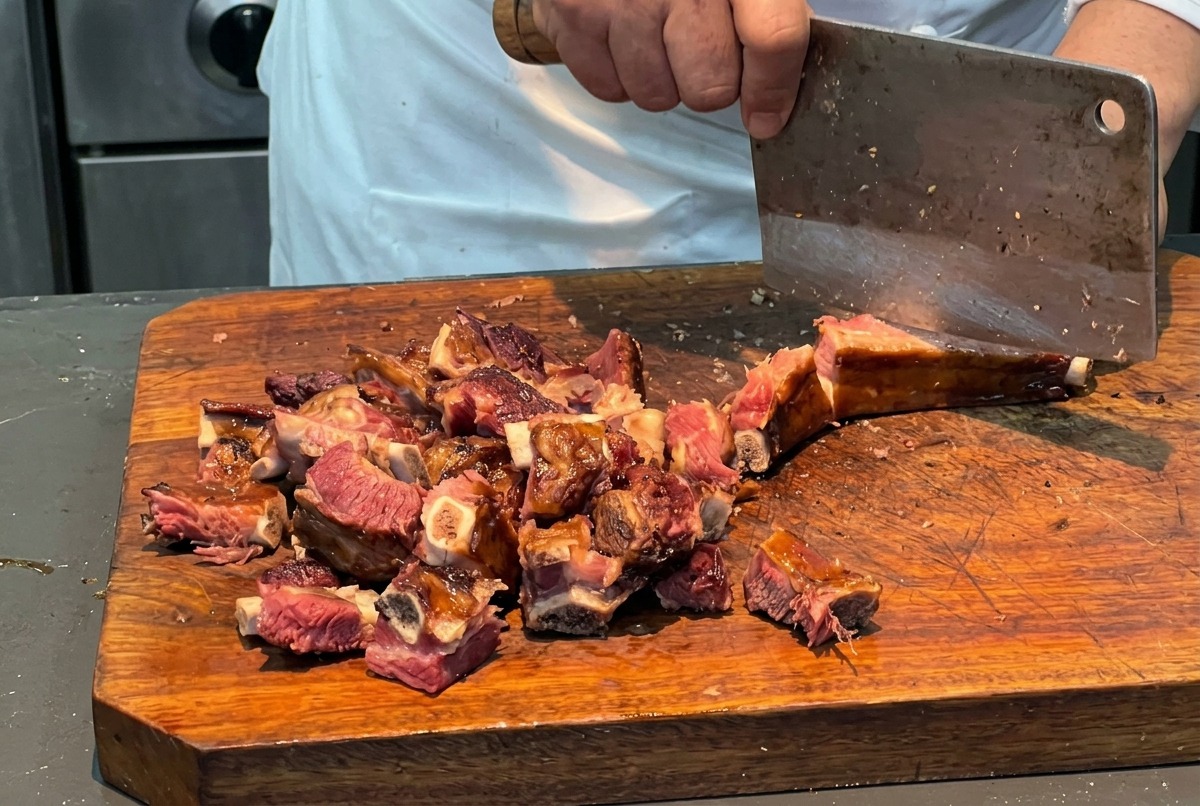 A chef using a large metal cleaver to chop a cooked slab of cured pork ribs into uniform 3-centimeter pieces on a thick wooden cutting board.