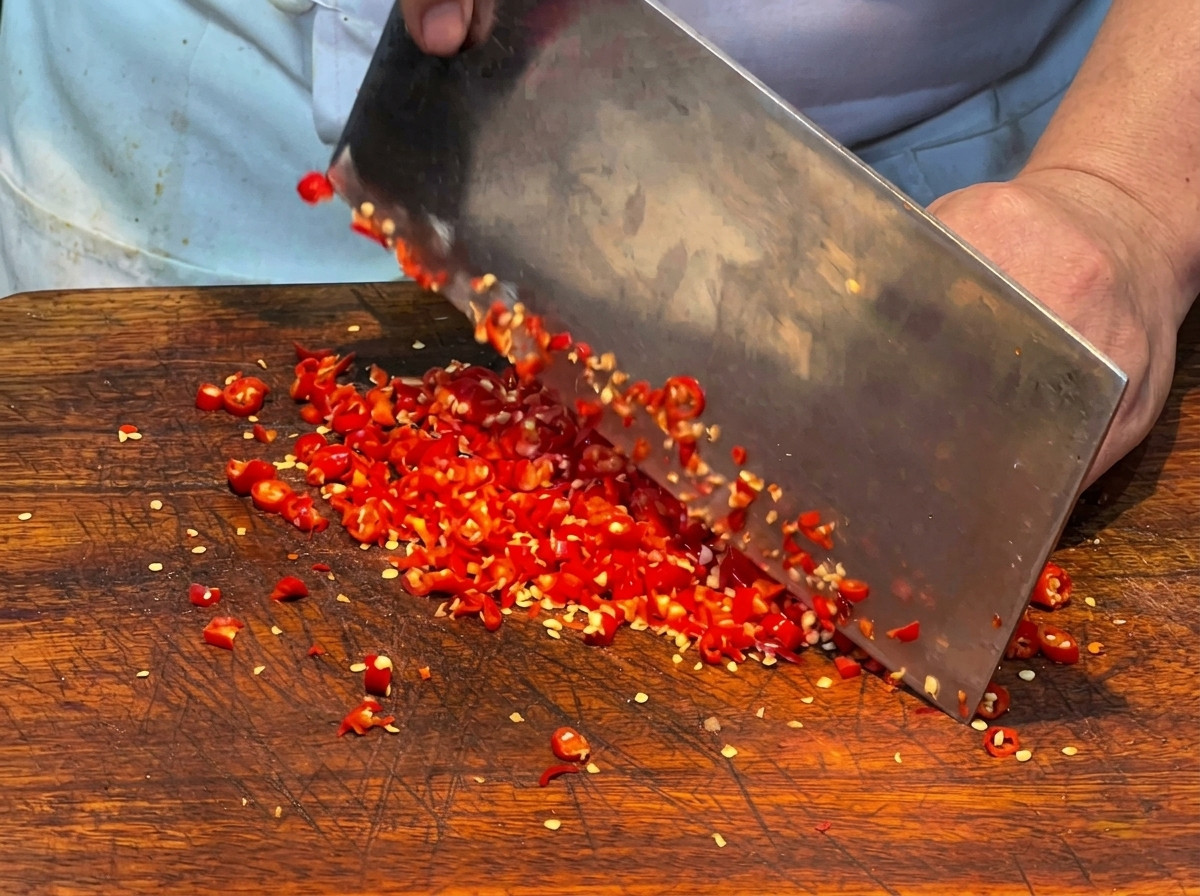 A close-up of fresh red chili peppers being finely chopped with a large metal cleaver on a wooden cutting board.