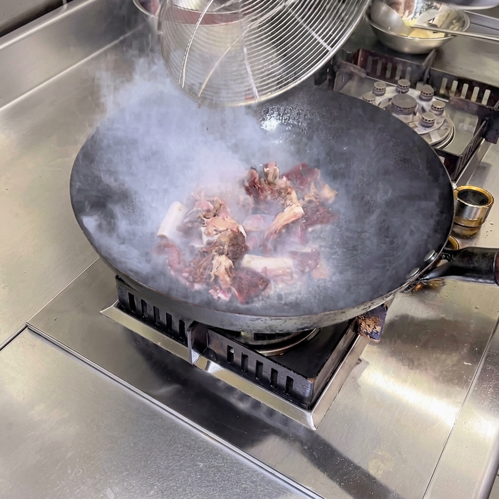 Blanched cured pork rib pieces being actively stir-fried over a high flame in a hot wok with camellia oil, with steam rising from the pan.