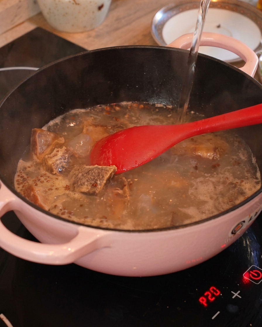 Hot water being poured into a pink cooking pot filled with fried cured pork ribs, ginger, and Sichuan peppercorns.