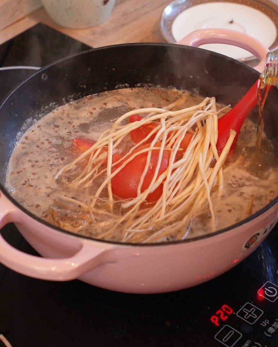 Tomato pieces and long white root herbs placed in a boiling pot with a clear broth, while cooking wine is being poured from a bottle.