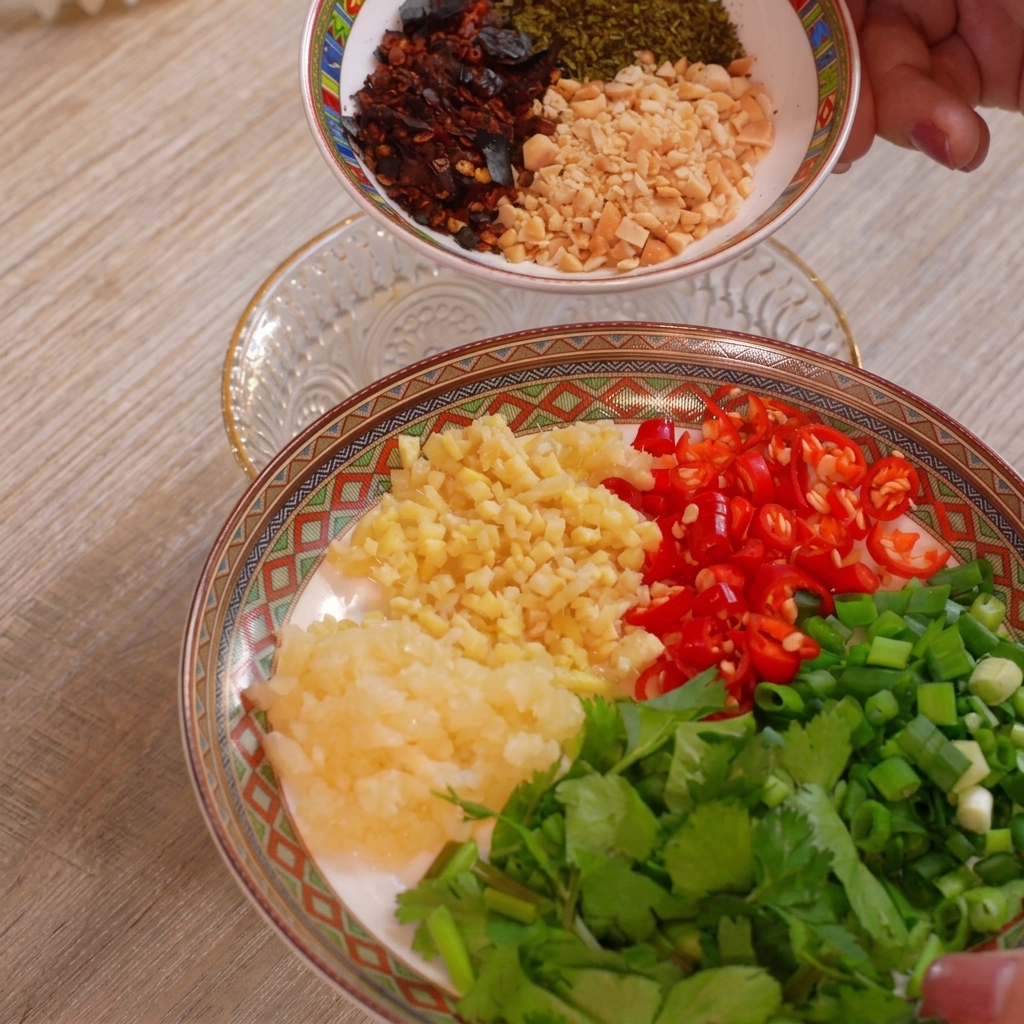 A plate arranged with separate mounds of minced garlic, minced ginger, chopped red chilies, green onions, and cilantro, with another bowl of peanuts and chili flakes held above.