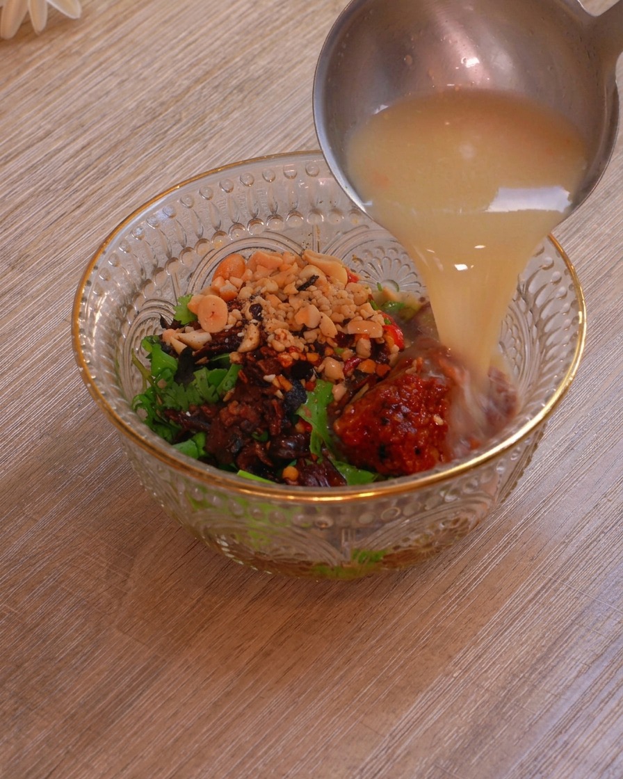 A metal ladle pouring rich, milky hot pot broth into a glass bowl layered with herbs, peanuts, chili flakes, and fermented bean curd.