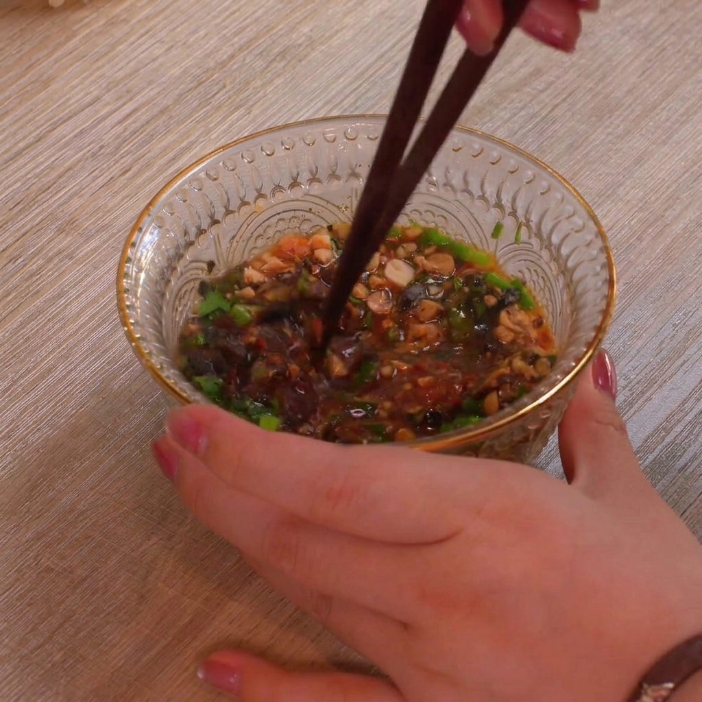 A close-up of a clear textured glass bowl filled with colorful dipping sauce ingredients like green scallions, crushed peanuts, and red chili flakes being mixed thoroughly with a pair of dark chopsticks.