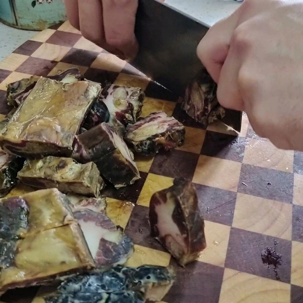 A close-up of a person using a large meat cleaver to chop cured pork ribs into small pieces on a wooden checkered cutting board.