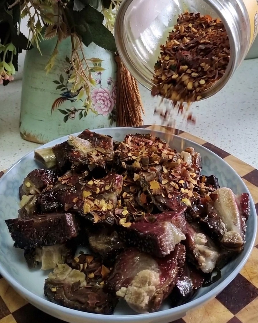 Red chili flakes being shaken from a glass jar onto a pile of cooked cured pork ribs on a plate.
