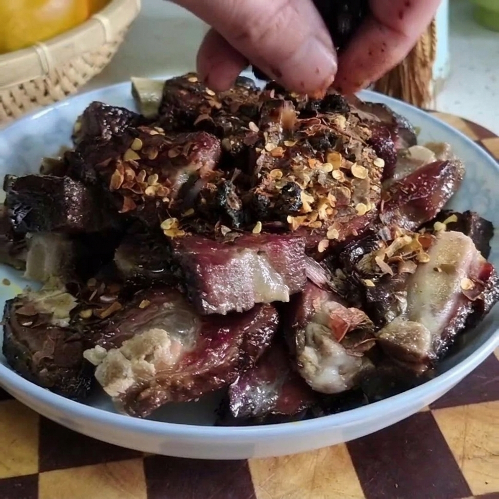 Close-up of a hand sprinkling fermented black beans over a plate of chopped cured pork ribs topped with chili flakes.