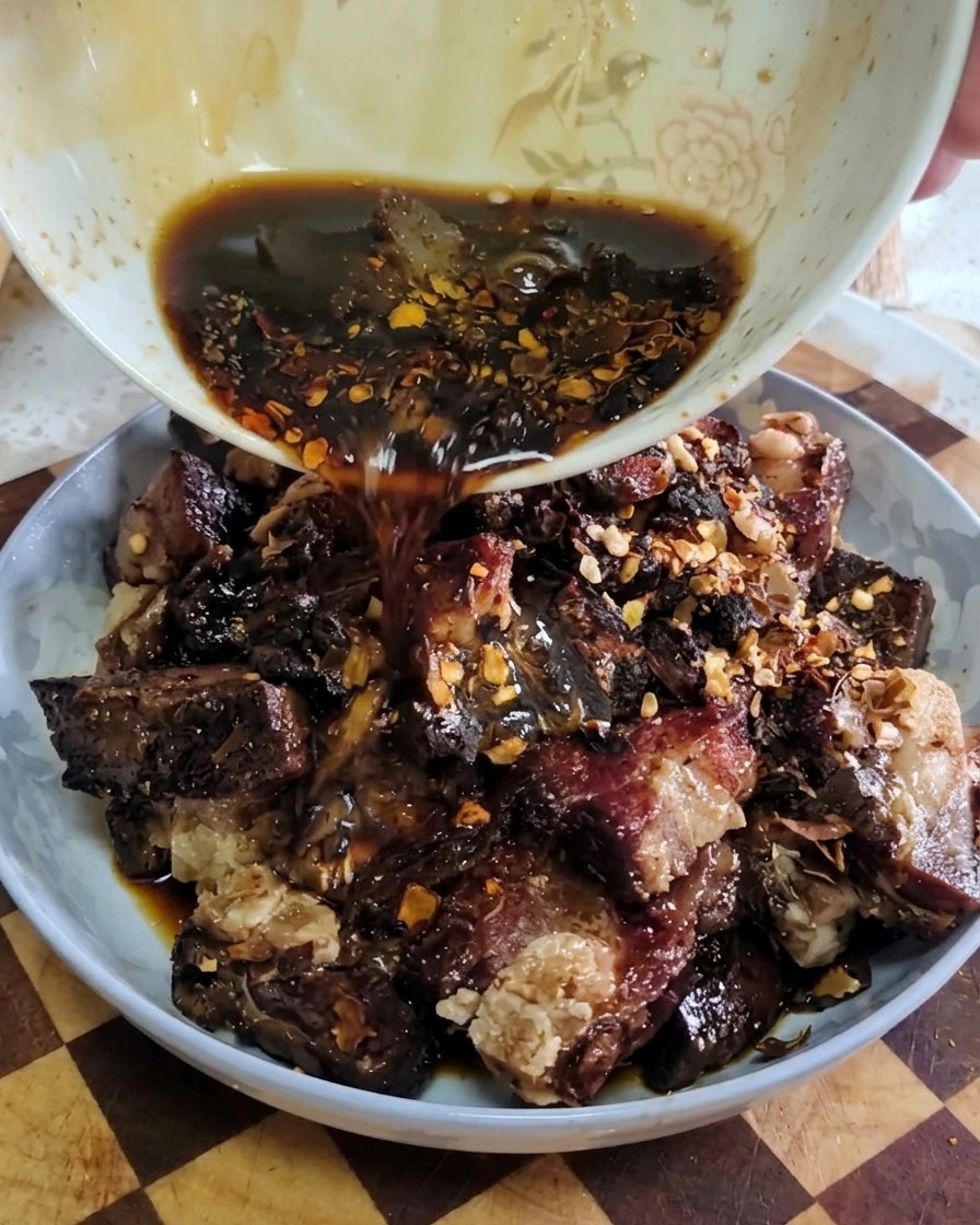 A dark savory liquid being poured from a small white bowl onto a plate of seasoned cured pork ribs.