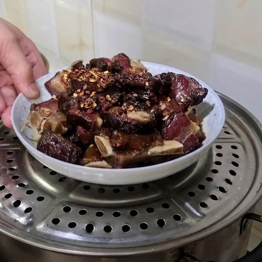A hand placing a plate of seasoned cured pork ribs onto a metal steamer rack inside a pot.