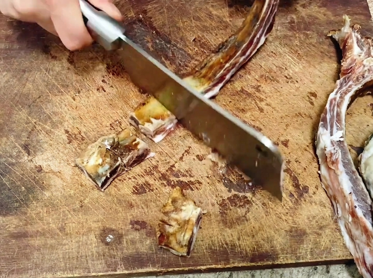 A large metal cleaver chopping a long cured pork rib into bite-sized pieces on a wooden cutting board.