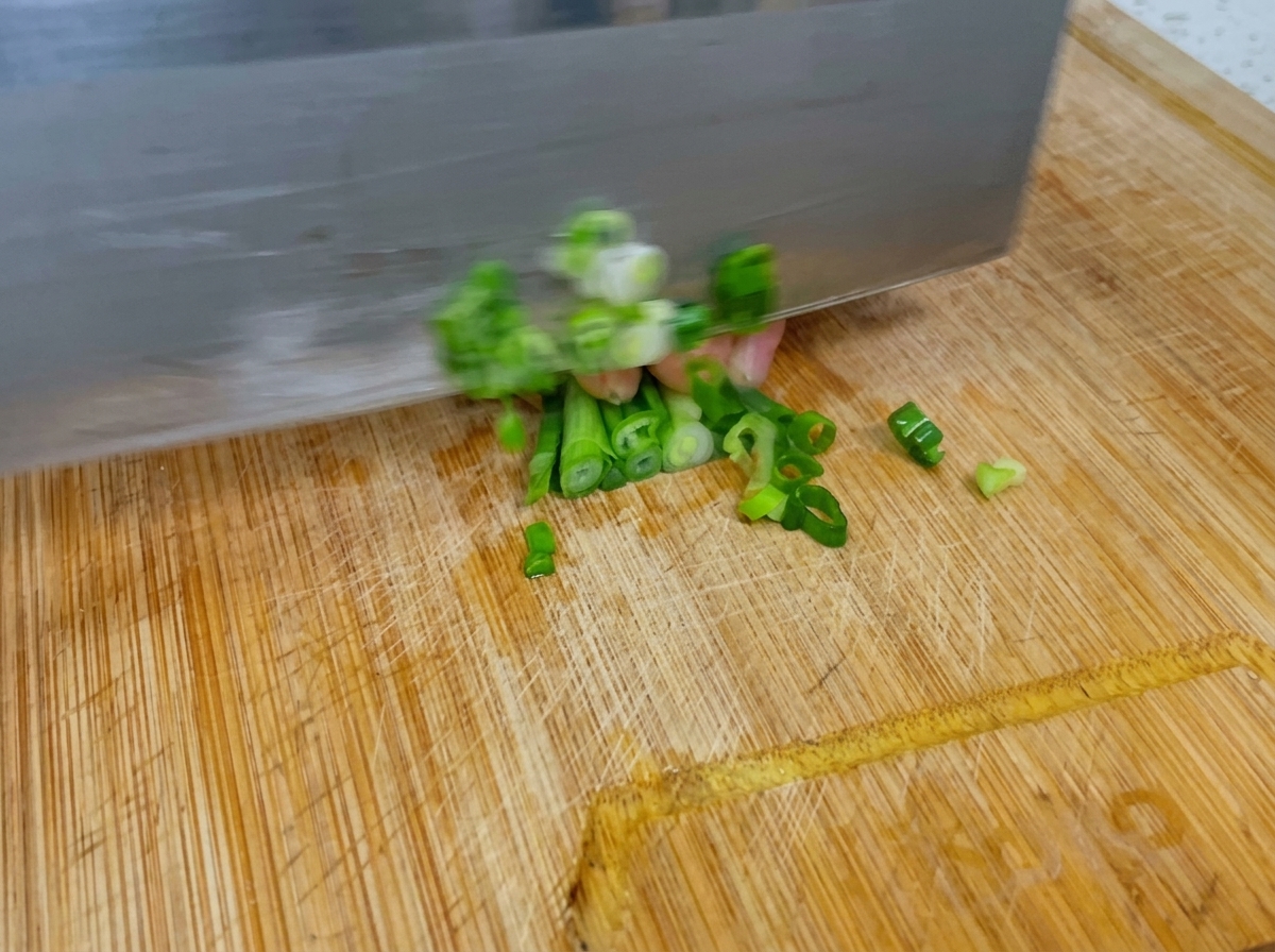 A knife quickly chopping fresh green scallions on a wooden cutting board.