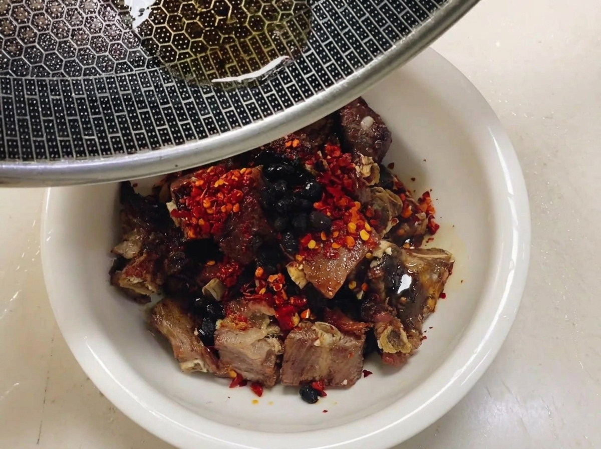 Pouring warm camellia oil from a honeycomb-textured metal wok over a seasoned bowl of pork ribs, red chili flakes, and black beans.