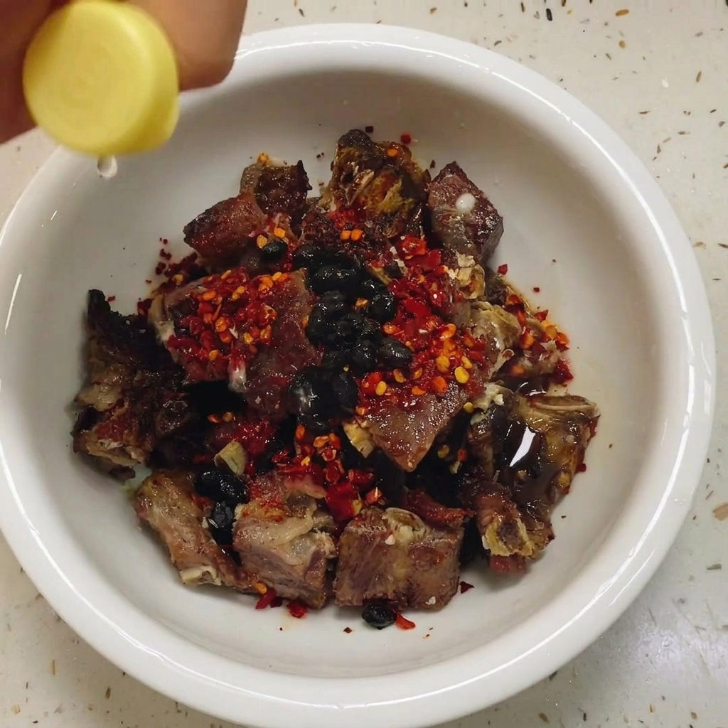 A hand adding a small splash of white vinegar from a bottle into a white bowl filled with seasoned cured pork ribs, chili flakes, and black beans.
