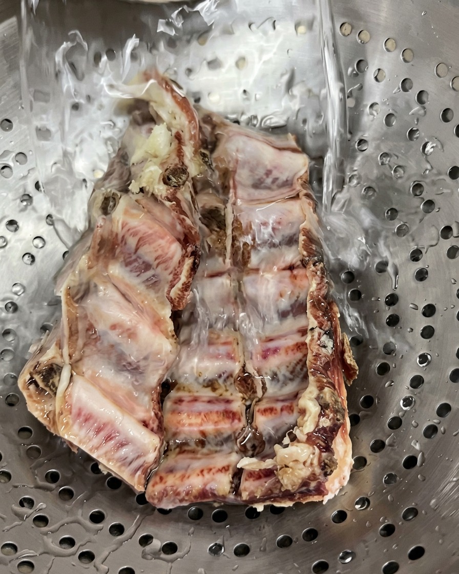 A raw slab of cured pork ribs sitting in a stainless steel colander being washed under a stream of clear running tap water.
