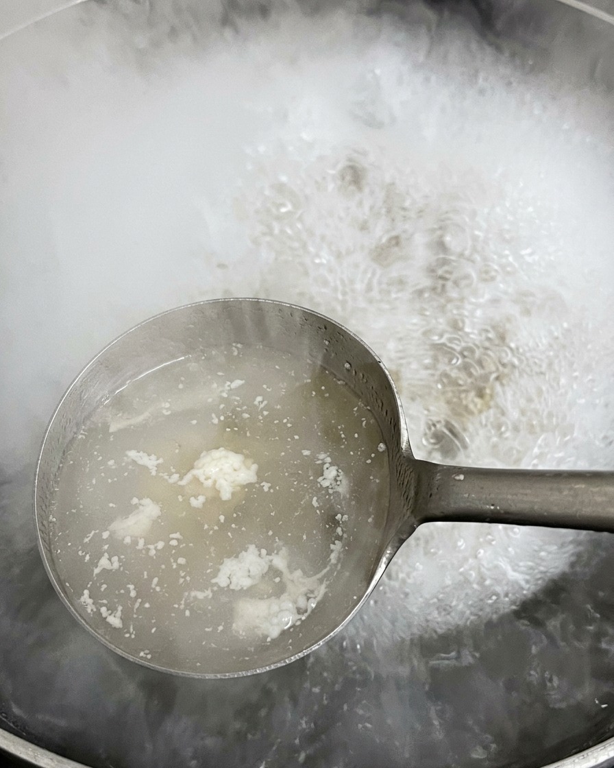 A metal ladle scooping up white frothy scum from the surface of rapidly bubbling and steaming hot water in a large pot.