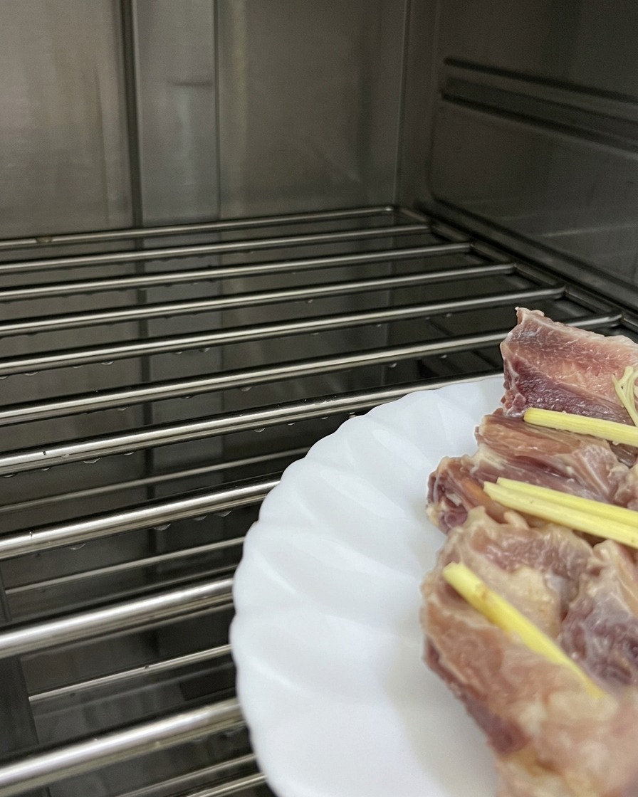 A white plate of pork ribs topped with ginger strips resting on a metal wire rack inside a steaming oven.