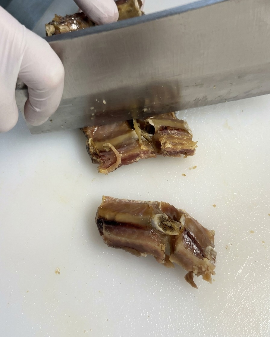 A chef wearing transparent gloves using a metal cleaver to chop cooked pork ribs on a white cutting board.