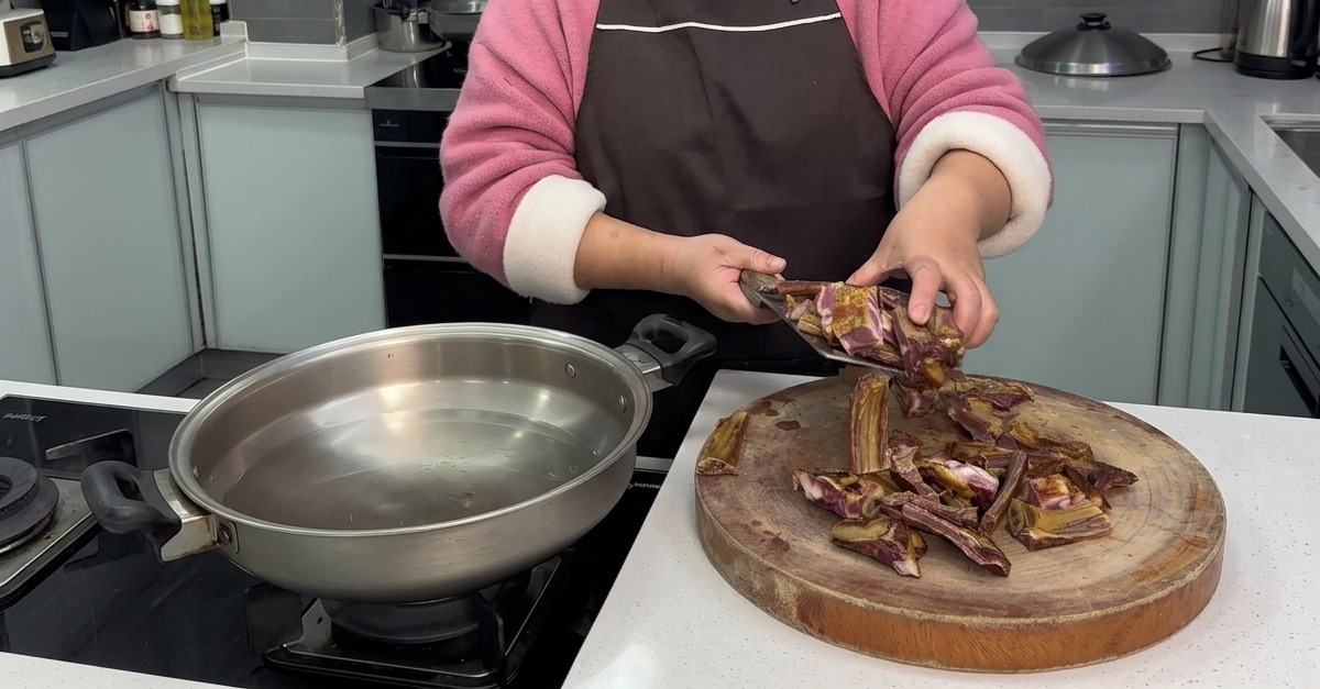 A person transferring freshly chopped pieces of cured pork ribs from a round wooden cutting board into a stainless steel pan of cold water.