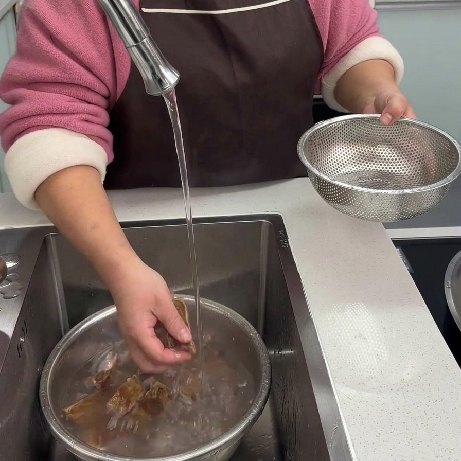 Hands thoroughly washing blanched cured pork rib pieces under a stream of clean running water in a kitchen sink bowl.