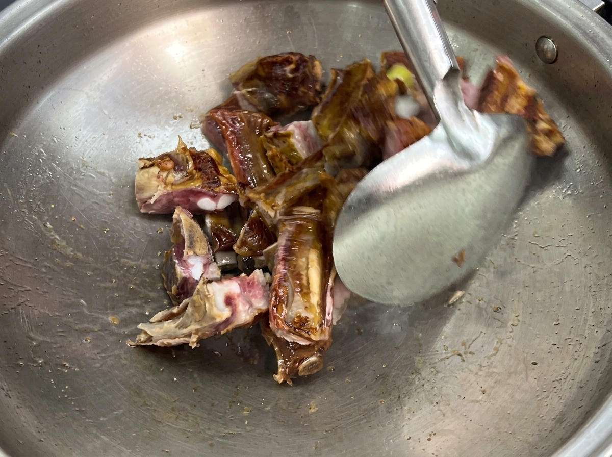 A metal spatula stir-frying pieces of cured pork ribs and sliced ginger inside a large stainless steel wok.