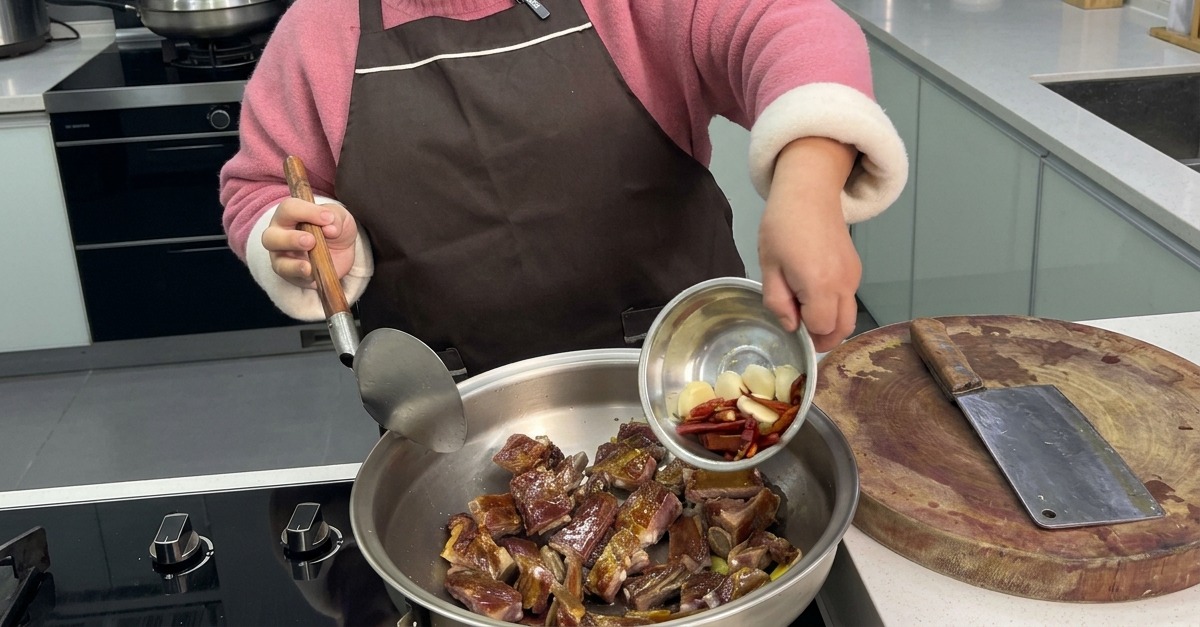 A person pouring whole garlic cloves and dried red chilies from a small metal bowl into a wok with stir-fried cured ribs.