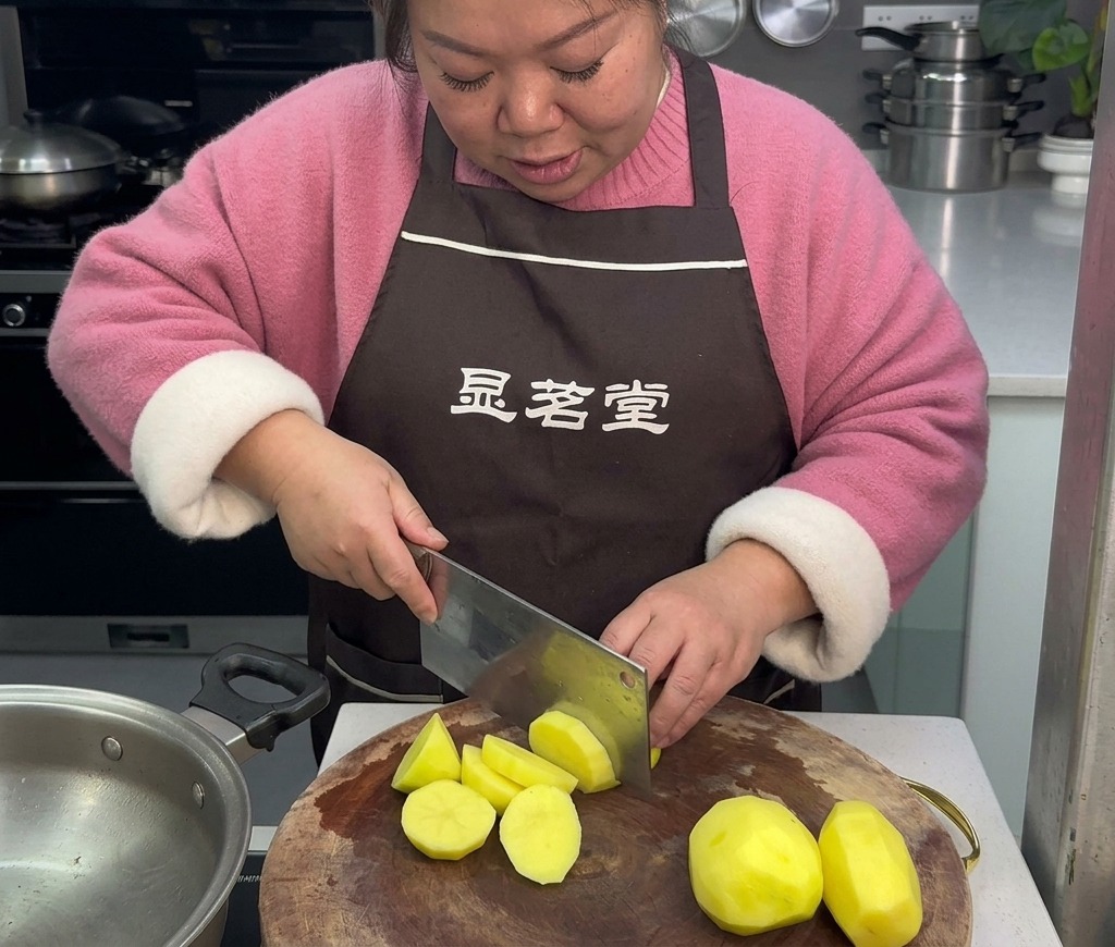 A woman wearing a brown apron uses a large kitchen knife to cut peeled yellow potatoes into roll-cut pieces on a round wooden cutting board.