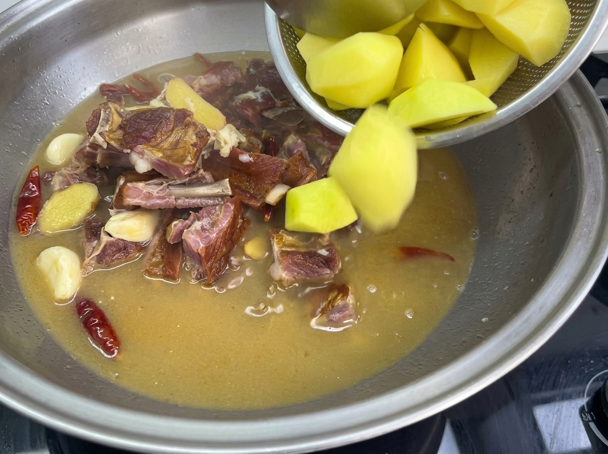 Pouring golden potato chunks from a mesh strainer into a large stainless steel pan filled with simmering broth, cured pork ribs, garlic, and red chilies.