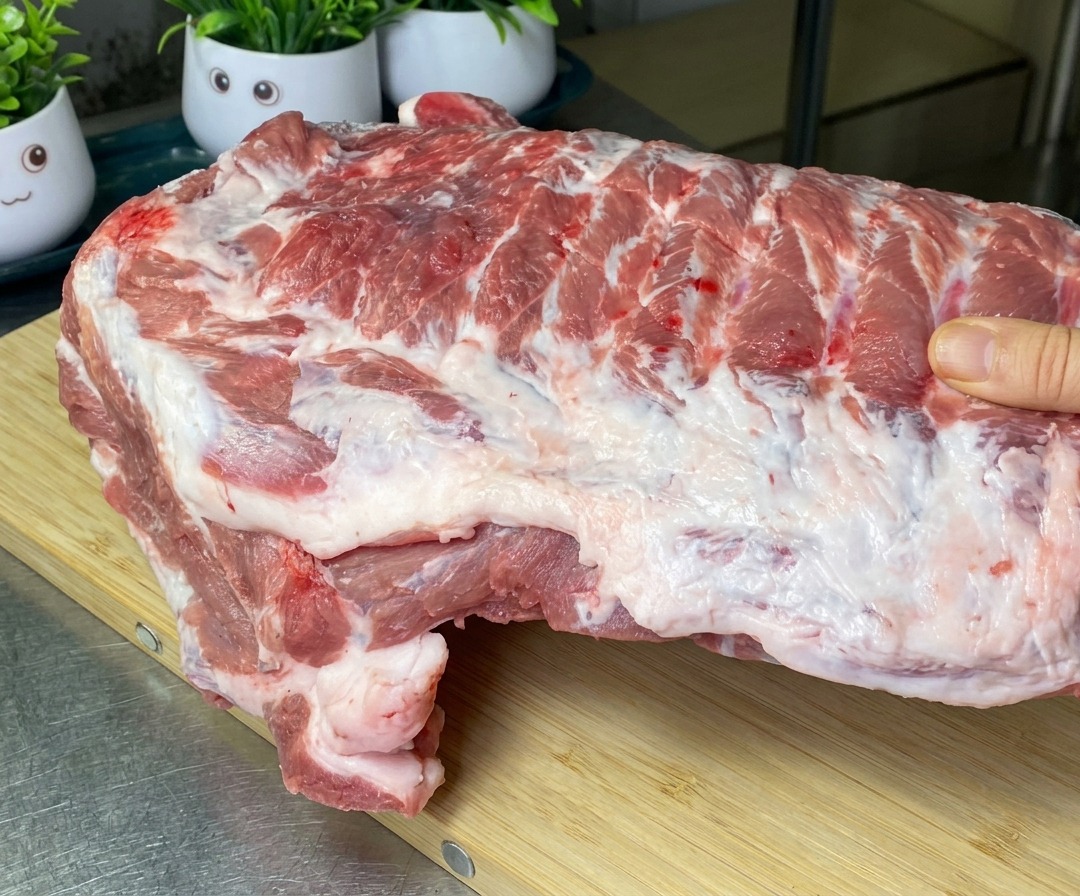 A large slab of raw pork ribs being held over a wooden cutting board, showing the initial cuts between the rib bones.