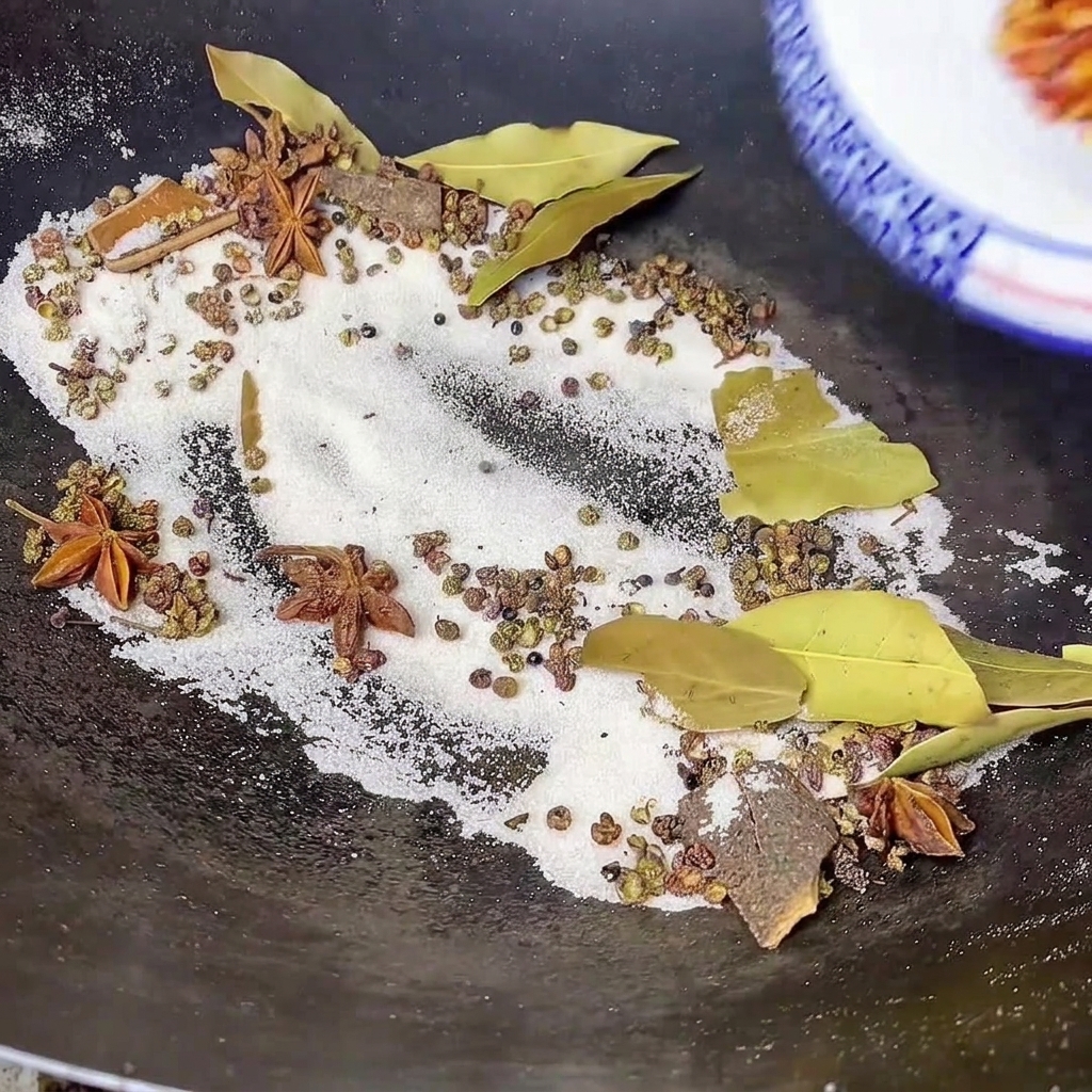 Red chili powder being poured from a bowl into a wok containing toasted salt, bay leaves, star anise, and other whole spices.