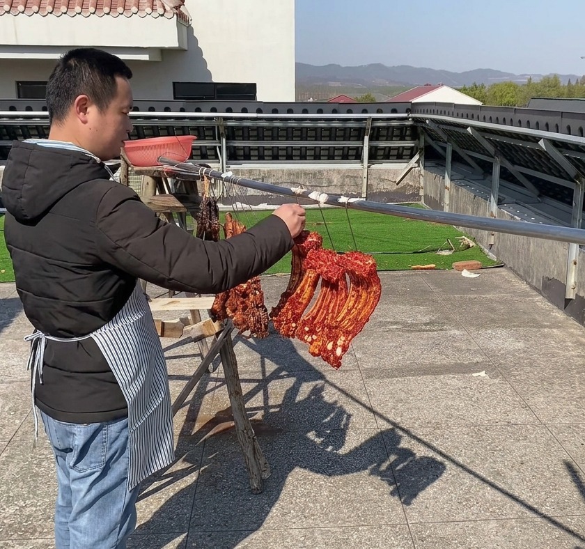Slabs of heavily spiced, deep-red pork ribs hanging side-by-side on a long outdoor metal bar to air-dry under a clear sky.